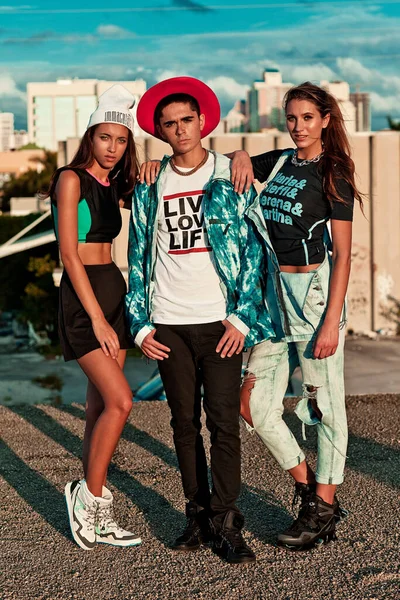 Portrait of an interracial group of 3 young adults, 1 male and 2 females, standing on top of a vacant building on a warm summer day wearing cool urban outfits.