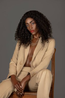 Portrait of a strong young black woman with long curly black hair and beautiful makeup sitting by herself on a wooden chair in a studio with a grey background wearing a beige suit and jewelry. clipart