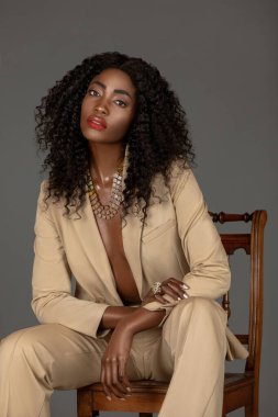 Portrait of a young black woman with long curly black hair and beautiful makeup sitting by herself on a wooden chair in a studio with a grey background wearing a beige suit and jewelry. clipart