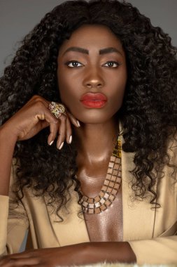 Uzun kıvırcık siyah saçlı, güzel makyajlı, gri bir arka planda, bej takım elbise ve mücevher giyen, tek başına kürkte poz veren, şehvetli siyahi bir kadının yakın plan portresi..