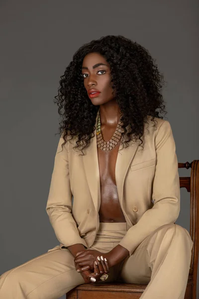 Portrait of an attractive young black woman with long curly black hair and beautiful makeup sitting by herself on a wooden chair in a studio with a grey background wearing a beige suit and jewelry.