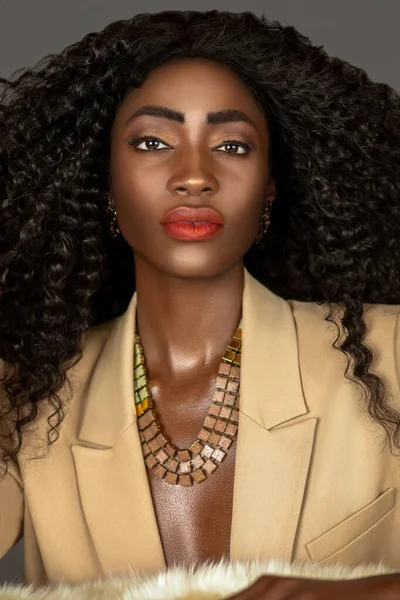 Closeup portrait of a strong black woman with long curly black hair and beautiful makeup posing by herself on a fur in a studio with gray background wearing a beige suit and jewelry.