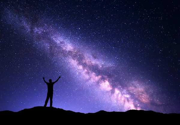 Colorido paisaje nocturno con púrpura vía Láctea y la silueta de un hombre deportivo de pie con los brazos levantados para arriba en la montaña. Cielo lleno de estrellas, espacio.. —  Fotos de Stock