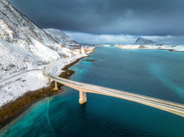 Norveç 'in Lofoten Adaları' ndaki turkuaz Arktik su üzerindeki bir köprünün havadan görüntüsü. Kışın karla kaplı dağlar, yollar ve kıyı şeridi. Manzaralı kuzey dramatik manzarası. Yol ve denizin en iyi drone görüntüsü
