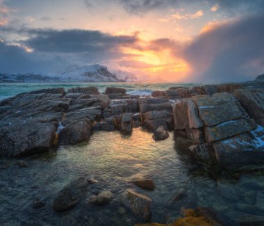 Kaya havuzları, kıyı taşları, kış dağları ve soğuk deniz suyu. Kutup manzarası, karlı kayalar, deniz, dalgalar, gün batımında dramatik bulutlu gökyüzü. Lofoten Adaları, Norveç 'te seyahat edin. Doğa. Deniz Burnu