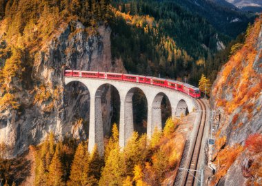 Alp dağlarındaki Landwasser viyadük üzerindeki modern kırmızı trenin havadan görünüşü, sonbaharda günbatımında renkli orman. Bernina Express, İsviçre. Sonbaharda tren, demiryolu, portakal ve kırmızı ağaçların üst manzarası