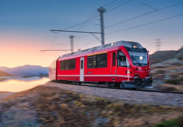 Sonbaharda günbatımında, dağ gölü yakınlarındaki modern kırmızı tren, karlı kayalar ve renkli gökyüzü. Bernina Express, İsviçre. Alplerdeki hız treni hareket bulanıklığı etkisi, sonbaharda suda yansıma