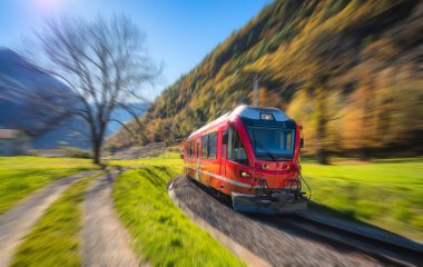 İsviçre 'deki güneşli sonbahar vadisinde yüksek hızlı kırmızı tren hareket ediyor. Renkli ağaçlar, yeşil çimenler, demiryolu rayları ve dağlar. Hareket bulanıklığı etkisi. Modern Bernina Ekspres treni. Bulanık arkaplan
