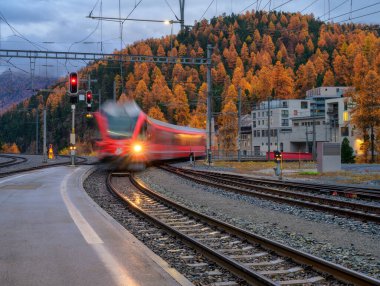 İsviçre Alpleri 'nde sonbahar yağmurunda dağ tren istasyonundan geçen bulanık kırmızı yolcu treni. Günbatımında hız treni hareket ediyor. St Moritz, İsviçre. Bernina Ekspresi. Demiryolu platformu ve ışıkları