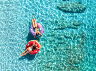 Mavi denizde yüzen iki güzel genç kadının hava manzarası. Yaz tatili. Açık turkuaz suda şişme halkaların üzerinde yüzen sportif kızların sırt üstü drone görüntüsü. Adriyatik Denizi, Hırvatistan