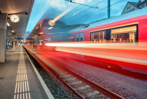 Bulanık kırmızı yolcu treni gece İsviçre Alpleri 'ndeki dağ tren istasyonundan geçiyor. Günbatımında hız treni hareket ediyor. St. Moritz, İsviçre. Bernina Ekspresi. Demiryolu platformu ve gece lambaları. Aktarım
