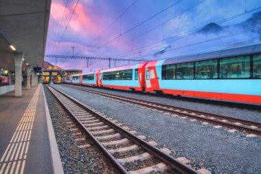 Alacakaranlıkta İsviçre Alpleri 'ndeki tren istasyonunda duran kırmızı ve mavi modern yolcu treni. Sis içindeki dağlar, raylar ve demiryolu platformu mor bulutlu gökyüzü altında. St. Moritz, İsviçre. Yüksek hızlı tren