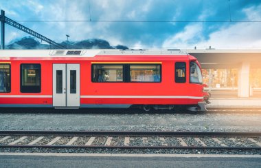 Sonbaharda İsviçre Alpleri 'ndeki dağ tren istasyonunda günbatımında kırmızı modern yolcu treni. Hızlı tren, demiryolu platformu ve bulutlu gökyüzü. Bernina Ekspresi. St. Moritz, İsviçre 'deki istasyon.