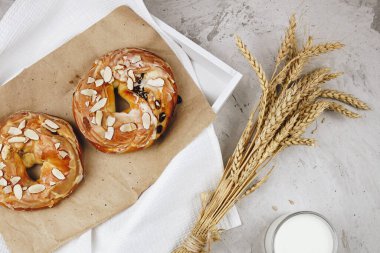 Kuru üzümlü ve bademli karamelli çörekler bir tepside kraft kağıdının üzerinde duruyor. Yanında bir buğday buğday ve çavdar kulağı, beton arka planda sütlü bir kupa, seçici bir odak.