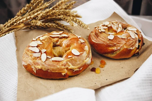 Kuru üzümlü ve bademli karamelli çörekler bir tepside kraft kağıdının üzerinde duruyor. Yanında bir buğday buğday ve çavdar kulağı var. Beton bir arka planda, seçici bir odak noktası..