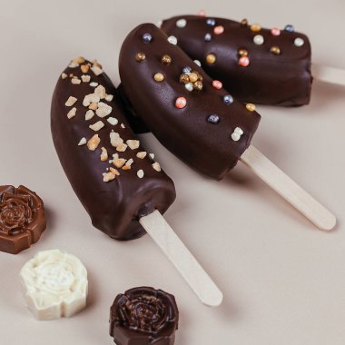 Üç tane çikolata kaplı muz, fındıkla süslenmiş ve bej arka planda serpiştirilmiş. Lezzetli tatlı. Yakınlarda çikolata var..