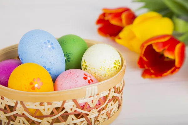 Coloridos huevos de Pascua en canasta de mimbre con tulipanes en madera blanca — Foto de Stock