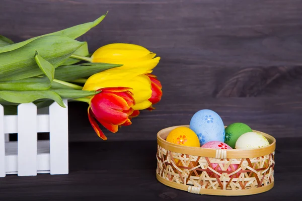 Colorate uova di Pasqua in cesto di vimini con tulipani su legno scuro — Foto Stock