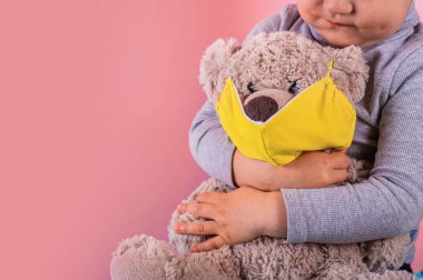 Küçük çocuk yüzüne sarı maske takmış oyuncak ayıya sarılıyor. Uzayı kopyala Bir salgın sırasında çocukları koruma kavramı.