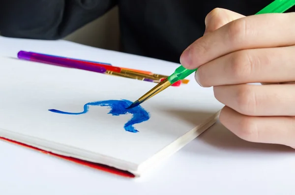 Left hand draws brush with blue paint on paper in album with sev