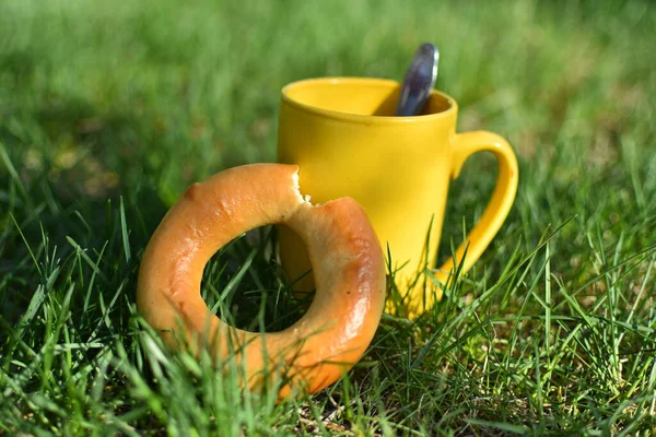 yellow mug stand on green grass. Nearby is a bagel. Photo taken with selective focus.