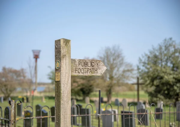 A wooden arrow sign post with Public Footpath on it and graveyard in background.