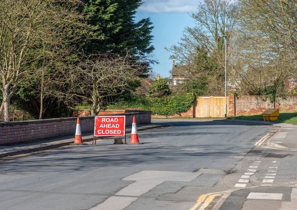 Red and white road closure sign with two red and white traffic cones in middle of street.