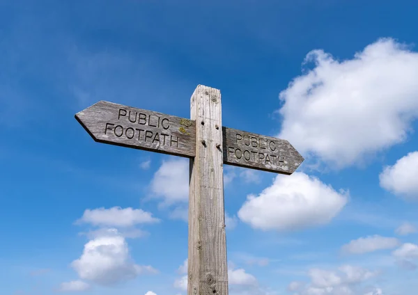 A double public footpath sign pointing in opposite directions on a wooden post with blue sky and clouds in the background.