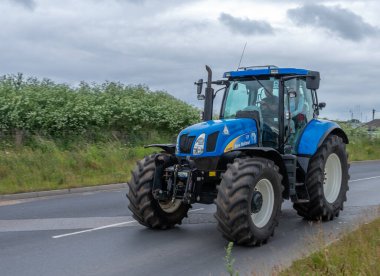 27 Haziran 2021 - Pocklington, Doğu Yorkshire, İngiltere - Beacon Genç Çiftçiler Kulübü Traktör Koşusu. Asfalt yolda çitli mavi traktör.