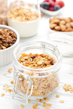 Cam kavanozda lezzetli granola ve beyaz ahşap arka planda kaseler.