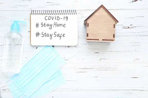 Inscriptions Stay Home, Stay Safe, Covid-19. Wooden house with medicine masks and sanitizer bottle on white background