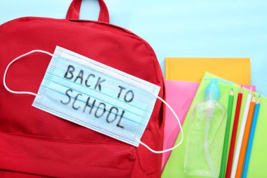 İlaç maskesi, dezenfektan şişesi, kırmızı sırt çantası ve mavi arka planda okul kırtasiyesi.