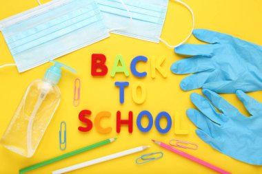 İlaç maskeleri, eldivenler, dezenfektan şişesi ve sarı arka planda okul kâğıdıyla okula dönüş.