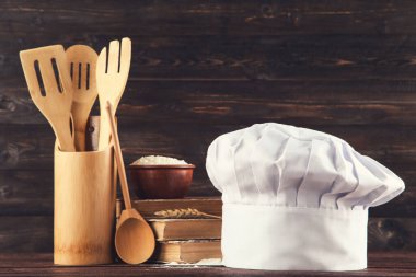 Kitaplı aşçı şapkası, bir kase un ve ahşap arka planda mutfak aletleri.