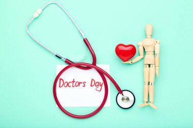 Kırmızı kalpli stetoskop, ahşap figür ve darphane arkaplanındaki Doktor Günü metni.