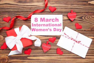 8 Mart Uluslararası Kadınlar Günü yazılı kart, kalpler ve zarf, hediye kutusu ve kahverengi ahşap arka planda kırmızı kurdele.