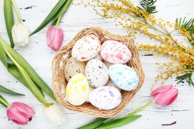 Beyaz ahşap arka planda lale ve mimoza çiçekli Paskalya yumurtaları sepette.