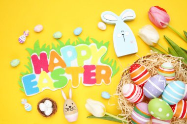 Mutlu Paskalyalar, renkli yumurtalar, ahşap tavşan ve sarı arka planda lale çiçekleri.