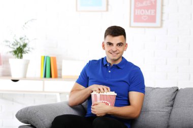 Elinde patlamış mısırla evdeki kanepede oturup film izleyen genç adam.