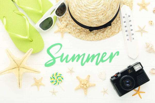 Text Summer with pair of flip flops, straw hat, seashells, thermometer and camera on white wooden background