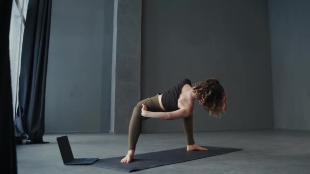 Ung attraktiv kvinna tränar gymnastiska stretching övningar med hjälp av sin dator. Online yogaträning koncept — Stockvideo