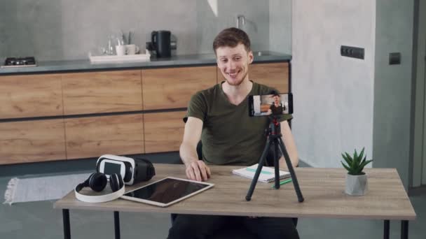 Young man blogger recording video blog using his mobile phone on tripod in the kitchen of his home. — Stock Video