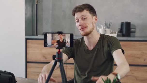 젊은 남자 블로거가 자기 집 부엌의 삼각대에 핸드폰을 올려 비디오 블로그를 녹화하고 있습니다.. — 비디오