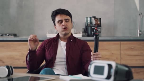 Ung man bloggare spela in video blogg med hjälp av sin mobiltelefon på stativ i köket i sitt hem. — Stockvideo