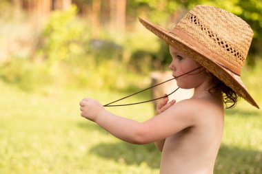 Hasır şapkalı küçük, odaklanmış bir çocuk boynundaki çizgiyi tutuyor ve uzaklığa bakıyor. Kırsalda harika çocukluk anları