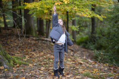 Sonbahar kıyafetleri içinde gülen tatlı bir kız sonbahar ormanındaki akçaağaç yapraklarına, doğadaki neşe ve çocukluk kavramına neşeyle uzanıyor..