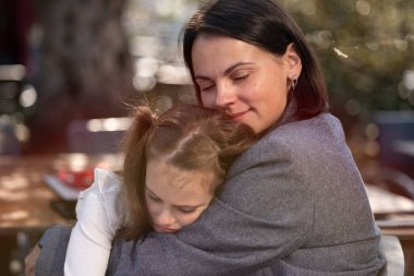 Bir kafede otururken yorgun kızını kucaklayan, sevgi ve duygusal destek kavramına sahip bir anne.