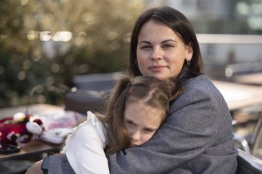 Gülümseyen anne yorgun kızına sarılıp yaz kafeteryasında oturmuş kameraya bakıyor. Aile sevgisi ve samimiyet kavramı.