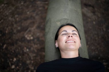 Siyah tişörtlü genç bir kadın ormanda devrilmiş bir ağaç gövdesine uzanmış hafifçe gülümsüyor yarı kapalı gözlerle, Almanya 'da doğal ışıkta yakın plan bir portre..