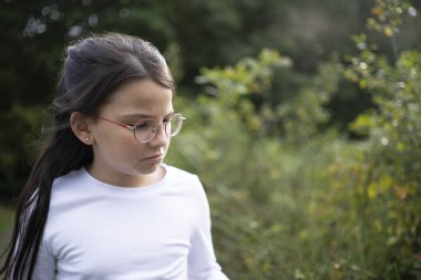 Gözlüklü, saçları arkadan bağlanmış bir kız ormanda dikiliyor, üzgün bir şekilde yan tarafa bakıyor. Yeşil çimenler, ağaçlar ve arka planda yumuşak doğal ışık, kopyalama alanı. Yalnızlık, yansıma ve duygusal hassasiyet kavramı..